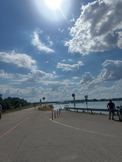 The image depicts a paved road leading towards a body of water under a bright, sunny sky with scattered clouds. On the left side of the road, there are trees and vegetation. The road has lane markings, including a pink line. Several traffic cones are visible along the side of the road. There are also traffic signs, one with a circular red border and a horizontal bar, and another with a white background and black text. On the right side of the road, a man wearing a black t-shirt and khaki shorts is standing and looking towards the water. A car is parked to his right. In the background, there is a body of water with several boats docked. The scene appears to be a waterfront area, potentially a marina or harbor, and is identified as being in Altenheim, Germany.
