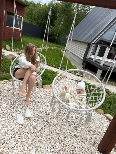 In Ryzhkovo, Russia, a young woman with long blonde hair sits in a white hanging chair swing, holding a disposable cup. She is wearing a white hoodie and dark shorts, with her legs crossed and bare feet dangling. Next to her, also in a white hanging chair swing, is an infant wearing a white hat and a matching cream-colored outfit, appearing to be a romper with a graphic design. The swings are suspended from a wooden structure. The ground is covered with white gravel, with patches of green grass and scattered rocks. In the background, a rustic wooden building and a modern A-frame house with a dark metal roof are visible. Lush green trees and vegetation surround the area. Two translucent white slippers are placed on the gravel near the woman's feet.