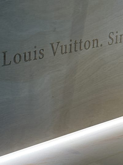 A close-up, slightly angled shot shows the words "Louis Vuitton. Sin." etched into a textured, light-colored surface. The text is in an elegant, serif font and is darker than the background. A bright, white light source runs diagonally across the bottom of the frame, illuminating a portion of the surface and creating a gradient effect. The overall impression is of a branded inscription on a material like wood or stone. The location in Nong Prue Subdistrict, Thailand, is not discernible from the image itself.
