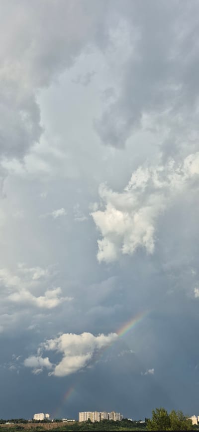 A cloudy, overcast sky with a faint rainbow is visible above a city skyline. The clouds are a mix of gray and white, with some darker storm clouds in the background. The rainbow is a thin arc of color, visible in the lower part of the frame. The city skyline consists of several apartment buildings and some trees. The image appears to be taken from a slightly elevated position, looking out over the city. The location is Codru, Moldova.