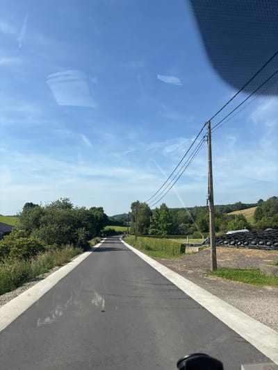 This media file displays an outdoor scene in Montzen, Belgium, on a clear, sunny day. The image is taken from a low angle, possibly from a moving vehicle, giving a dynamic perspective of a paved road extending into the distance.

The foreground is dominated by the road, which has a white line marking its edge. To the left of the road, there is a clear blue sky with scattered wispy clouds, and a reflection of what appears to be a textured surface, possibly from the vehicle's interior, is visible on the sky. Further to the left, a portion of a dark, patterned object occupies the edge of the frame.

A wooden utility pole with several wires stretches across the frame, positioned between the sky and the road. Beyond the pole, the landscape unfolds with rolling green hills covered in lush trees and fields. On the right side of the road, a stack of dark, circular objects, resembling tires or large rings, is visible against a backdrop of gravel and green grass. The scene suggests a rural or semi-rural setting, with evidence of infrastructure like the utility pole and the paved road.

The sunlight indicates it is daytime, likely mid-morning or afternoon. The clear sky and greenery suggest pleasant weather. There is no visible text in the image. The overall impression is one of a serene and open landscape on a bright day.