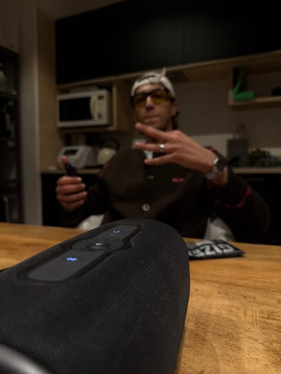 The image captures an indoor scene, likely a kitchen or dining area within a residence in Saint-Denis, France, with a primary focus on a foreground object. A black, cylindrical portable speaker, featuring a textured fabric exterior and a control panel with illuminated blue icons, is positioned on a light brown wooden table, in sharp focus.

In the blurred background, a male individual is seated. He wears yellow-tinted glasses, a white head covering (possibly a cap or bandana), and a dark brown jacket with lighter-colored trim. His left hand holds a dark-colored smartphone. His right hand is raised, displaying a "V" gesture (peace or victory sign), with a silver-toned ring visible on his right ring finger and a watch on his right wrist.

The environment behind the individual includes dark wall-mounted cabinets, a light-colored microwave oven, and a white kettle on a countertop. A dark, rectangular object, possibly a decorative item or a license plate with partial white lettering ("SIZE"), rests on the table surface behind the speaker to the right. The scene is illuminated by interior lighting.