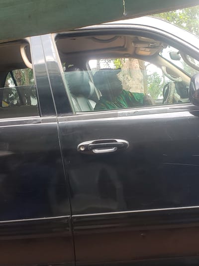 The image shows the side of a black vehicle, possibly a car or truck, with an open door. The door handle is visible. Through the window, part of the interior of the car is visible, including seats and the steering wheel. The interior appears cluttered with a green and yellow patterned fabric draped over the steering wheel. The background shows a blur of trees and vegetation. The photo was taken in the city of Umundugba, Nigeria.