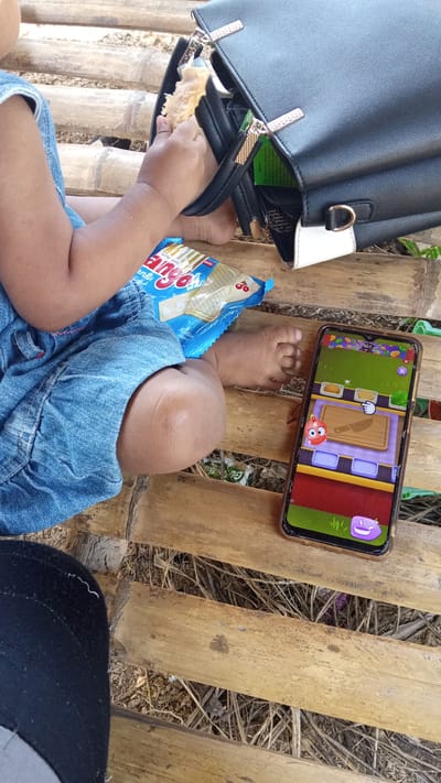 A close-up shot shows a child's hands and legs resting on a bamboo surface. The child is wearing a blue denim dress and is holding a piece of food from a bag of snacks. A smartphone is placed next to the child, displaying a game interface. A black handbag is also visible in the background. The environment appears to be outdoors, possibly in a rural setting, with dry grass and soil visible between the bamboo slats. The image is taken in Meunasahdayah, Indonesia.