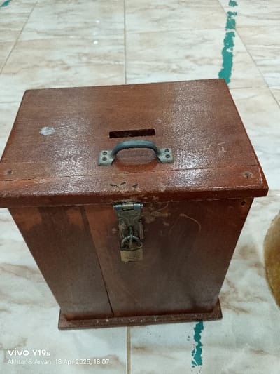 The image shows a wooden box with a metal handle on the top and a metal lock on the side. The box is brown and has a slightly worn appearance. The image appears to be taken indoors on a tiled floor with green grout. The location, based on the provided coordinates, is a residential area in Kecamatan Kroya, Indonesia. The image depicts the box with no people or other objects in the scene. The box is closed, and the lock is in place, indicating that the box is secured. There are no indications of any actions, interactions or other noteworthy details.