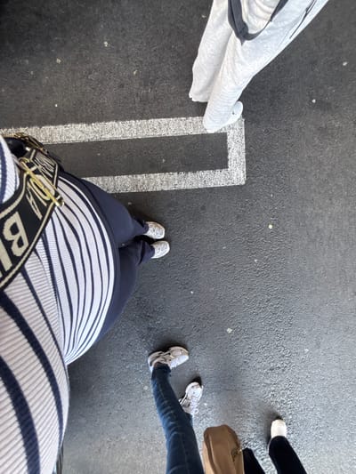 The scene depicts an overhead view of four individuals standing on dark grey asphalt pavement featuring a white painted rectangular ground marking. The precise location within Culver City, United States of America, is not identifiable from the visual content.
One individual, positioned on the lower left, wears a white and navy blue striped garment, navy blue pants, and light-colored athletic shoes. A black and white strap with gold-colored hardware and partial "BI" lettering is visible across their torso. Their feet are positioned adjacent to the left side of the white rectangular marking.
To the top right, a second individual's lower body is visible, wearing light grey sweatpants and white athletic shoes with a visible black Nike swoosh. Their right foot is placed inside the white rectangular line. A black strap is visible over their shoulder.
A third individual is located in the bottom-right quadrant, wearing blue jeans and white athletic shoes. A tan handbag rests on the ground near their feet.
The lower legs and white athletic shoes of a fourth individual, wearing dark pants, are partially visible at the far bottom-right.
All subjects are standing. No explicit actions or interactions beyond their shared presence on the pavement are depicted. The asphalt surface shows minor texture and scattered light-colored debris.