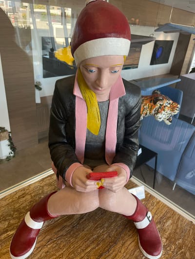 This image presents a brightly painted, full-body sculpture of a blonde female figure, positioned in a seated posture on a polished, light brown surface with a marbled pattern. The figure wears a red Santa hat with a white brim, a black blazer featuring pink lapels, a gray shirt, and red knee-high boots with white cuffs. Her light blue eyes are directed downward, focusing intently on a small, red, rectangular object held in both hands, resembling a smartphone, with yellow polish visible on her thumbs.
The background, viewed through a reflective glass surface, reveals an indoor commercial or gallery setting located in Galatina, Italy. Visible environmental elements include light blue upholstered chairs, a dark table, and various wall-mounted artworks, specifically a large yellow abstract painting, a dark blue circular piece, and a black and white landscape. A mosaic-style sculpture of a tiger's head is prominently displayed on or near a blue chair. Additionally, small models, including a light blue airplane, are suspended from the ceiling. A white informational label is situated on the surface to the right of the sculpture's knee.