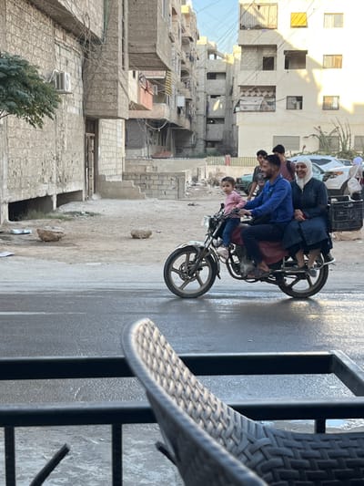 The image depicts an urban street scene in Mu'adammiyat ash-Sham, Syria, captured from a slightly elevated perspective.
In the midground, a family of three rides a dark-colored motorcycle from left to right on a wet or reflective asphalt road. The driver is an adult male wearing a blue long-sleeved shirt, dark pants, and sandals, smiling and looking slightly away from the camera. A young child, dressed in a pink long-sleeved top and dark pants, sits directly in front of the driver, facing forward with a neutral expression, looking towards the camera. An adult female, wearing a white hijab and a dark blue dress, sits behind the driver, also in sandals, smiling and looking slightly away from the camera. A black plastic crate is attached to the rear of the motorcycle.
The background features a dirt and gravel shoulder adjacent to the road, littered with a few rocks. Beyond this, several multi-story residential buildings, primarily constructed of light-colored concrete blocks, line the street. These buildings exhibit visible utility lines, exposed rebar in some areas, and what appear to be weathered or incomplete finishes. An outdoor air conditioning unit is visible on a building to the far left, where a green tree also stands. Further back on the right, parked vehicles, including a white car, are visible. Several individuals are present in the background: two younger males stand near the parked cars, and an additional person in a white head covering is seen near a vehicle. The sky above is clear and light blue, indicating daylight.
In the immediate foreground, the top of a light-colored woven outdoor chair and a black metal railing are partially visible, suggesting the photograph was taken from a balcony or outdoor seating area.