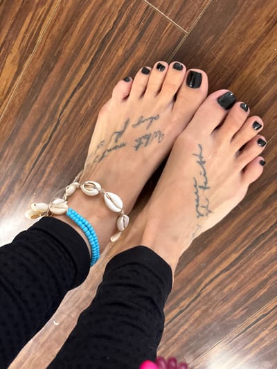 This full-shot image features a person's bare feet resting on a polished wooden floor. Both feet have glossy black nail polish applied to the toenails. Script tattoos are visible on the top of each foot. The tattoo on the left foot appears to read "What meant always...", while the tattoo on the right foot is less clear but seems to say "Your Faith".
The right ankle is adorned with two anklets. The prominent one is made of natural cowrie shells strung together on a cord. Beneath this, a second anklet consists of multiple strands of small, light blue beads. Black fabric, likely from clothing such as leggings or trousers, is visible around the lower calves. A small, out-of-focus pink or purple object is partially visible on the far right edge of the frame. The background consists of a wooden floor with varying shades of brown planks, suggesting an indoor setting. The image, set in Seal Beach, United States of America, focuses on showcasing the foot adornments and tattoos.