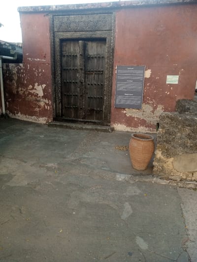 A weathered, reddish-brown plastered building facade, exhibiting significant peeling, features a centrally located, intricately carved dark wooden double door. The door and its frame display detailed Swahili-style carvings, and the panels are adorned with circular metal studs. To the left of the door, a white PVC downspout runs vertically. To the right, a dark grey rectangular information sign, partially showing "Munge at Msuri" and "Mungwana wa Nasari" below Arabic script, is affixed to the wall; a smaller, lighter sign is adjacent. A ribbed, unglazed terracotta pot, light brown in color, rests on the uneven, cracked grey concrete ground to the right of the door, beside a rough-hewn stone structure. The ground contains scattered organic debris. No people or active interactions are depicted. This scene is located in the Mji Wa Kale (Old Town) area of Mombasa, Makadara ward, Kenya.