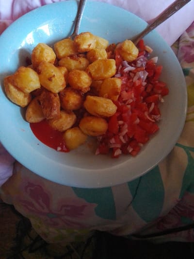 A light blue bowl occupies the center of the frame, containing a meal. On the left side of the bowl are numerous golden-brown, irregularly shaped fried or roasted potato pieces. On the right side, there is a mound of finely diced red tomatoes mixed with white and light purple onion pieces. A small puddle of red sauce, likely ketchup, is visible on the bottom left of the bowl, partially covered by some potato pieces. Two silver-colored spoons, featuring ornate handles, are partially visible within the bowl's contents. The bowl is positioned on a multi-patterned fabric surface, which includes light blue striped sections and large pink floral designs on a light background. No individuals are present. The scene is located in Karingani ward, Kenya.