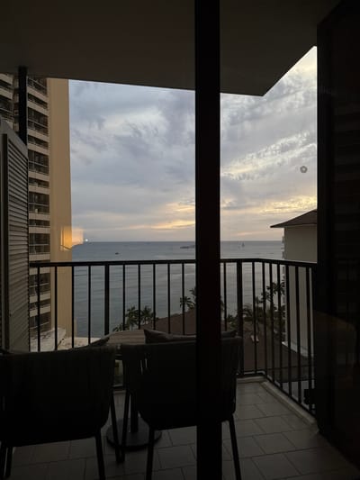 A slightly rotated, medium-shot, indoor photograph from a high vantage point looks out towards the coast. A set of patio chairs and table sit at the lower-right of the view, partially blocking the ocean view. A set of palm trees are visible between the patio railings on the right. The ocean is in the center-right of the shot. A building can be seen on the lower-left and in the center, below the view of the ocean. The sky is in the upper-left of the frame, and a parasail can be seen towards the upper-left. The location is Honolulu, Hawaii.