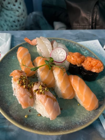 An overhead shot captures a visually appealing plate of assorted sushi, presented in an indoor setting. The main subject is a rustic-looking, teal-green ceramic plate filled with various types of nigiri and gunkan maki.

On the plate, there are several pieces of salmon nigiri, featuring slices of light orange salmon draped over white rice. Two pieces of gunkan maki are visible, consisting of rice wrapped in dark seaweed and topped with bright orange, finely chopped salmon or salmon roe. Further pieces include what appear to be shrimp or squid nigiri, characterized by translucent white toppings over rice, one piece clearly showing a pink shrimp tail. Additionally, two pieces of salmon nigiri are garnished with a sprinkle of chopped green pistachios and a light drizzle of sauce. The sushi is artfully arranged and accompanied by a thin slice of red radish and a sprig of fresh green pea shoots, adding vibrant accents to the dish.

In the blurred background on the left, a dark leather jacket is partially visible, suggesting a casual dining environment. Towards the bottom left, a light-colored textile with a brown spotted pattern is also visible. In the upper right corner, a piece of paper with some faint, illegible text and a clear QR code is present, possibly a receipt or a menu. The overall scene suggests a meal being enjoyed in Basiglio, Italy, during an unspecified time of day, with soft ambient lighting.