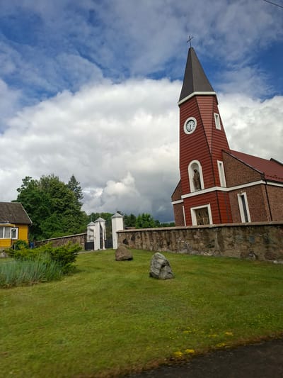 The image depicts the Church of St. Joseph in Balbieriškis, Lithuania. The church features a prominent red brick facade and a distinctive red tower with a dark roof, topped by a cross. A clock face is visible on the tower, along with a niche containing a statue. A stone wall with a gate separates the church grounds from the surrounding area, which includes a yellow house with a grey roof and green trees. The scene is set against a backdrop of a dynamic sky with both blue patches and large, white clouds, some of which appear dark and heavy, suggesting potential weather changes. The foreground is a lush green lawn with scattered yellow wildflowers and a few large rocks.