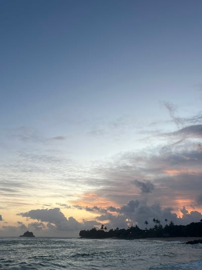This is a landscape photograph of a beach scene at sunset in Midigama, Sri Lanka. The image is vertically oriented.

The scene depicts a tropical coastline with the ocean on the right and a densely vegetated, hilly shoreline on the left. The ocean waves are visible, appearing textured and grey. The shoreline is characterized by dark green foliage, with several tall palm trees silhouetted against the colorful sky. A small, dark rocky outcrop is visible in the water near the shore.

The sky is a dominant feature, displaying a dramatic sunset with a mix of clouds. The left side of the sky is a soft, clear blue, transitioning to lighter shades with wispy white clouds towards the center. Further to the right, the clouds are denser and darker, interspersed with vibrant streaks of orange, pink, and yellow from the setting sun. This gradient of color suggests the time of day is either sunrise or sunset.

There are no people or discernible objects other than natural elements. The overall mood evoked is serene and tranquil, typical of a peaceful beach sunset. No text is visible in the image.