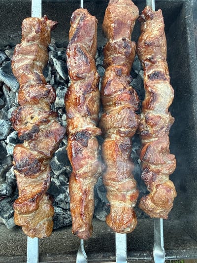 The image shows four metal skewers holding grilled meat, likely shish kebab, positioned over a bed of hot charcoal in a grill. The meat appears browned and cooked, with some areas darker than others. The grill's surface is visible around the charcoal. There is no indication of the location, Granovshchina, Russia, or the presence of any people.
