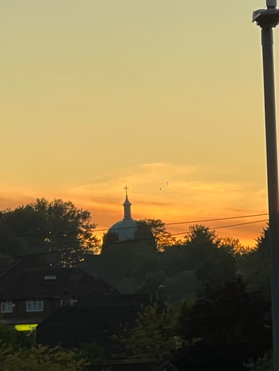 The image, rotated 90 degrees clockwise, depicts a tranquil twilight scene over High Wycombe, United Kingdom, likely captured during either sunset or sunrise. The expansive sky, occupying the left and upper portions of the frame in this orientation, showcases a beautiful gradient of warm colors, moving from a soft pale yellow-orange on the far left to richer hues of orange and pinkish-gold towards the center and right. Wispy clouds, illuminated by the low sun, add texture to the atmospheric display.
Along the right side and extending into the bottom-right, a dark silhouette of a tree line and dense foliage forms a natural boundary against the luminous sky. Partially emerging from behind these trees, and silhouetted against the brightest part of the sky, is the distinctive dome or cupola of a church, topped with a prominent cross. The church's structure appears mostly dark against the vibrant backdrop.
In the bottom-right corner, a section of a brick building with visible windows is discernible, largely in shadow and partially obscured by the surrounding trees. A thick, dark pole or beam runs horizontally across the top-left corner of the image. Two very small, distinct colored specks, one pink and one greenish-blue, are visible in the upper-middle sky, above and to the left of the church, possibly indicating distant hot air balloons or atmospheric phenomena. No discernible text is visible.