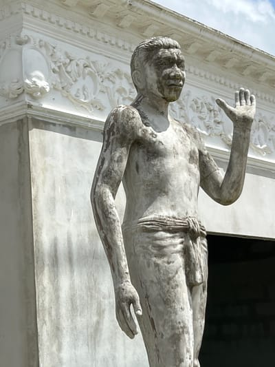 The image shows a weathered, pale gray statue of a standing male figure in what appears to be an outdoor setting in Pa Khlok, Thailand. The statue depicts a man with a broad chest and defined musculature, wearing a simple sash around his waist and lower torso. His right arm is raised, with an open palm facing forward. The statue's surface shows signs of age and wear, with patches of darker discoloration and a rough texture.
Behind the statue, a building with white, ornate architectural details is visible. These details include intricate moldings and decorative carvings, suggesting a temple or a historically significant structure. The building's wall to the left of the statue is a plain, textured gray. The sky above is partly cloudy with a mix of white clouds and blue sky. The overall impression is one of historical preservation and reverence.