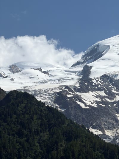 A high-angle, vertical medium shot captures a majestic mountain landscape under a clear blue sky with scattered white clouds. The foreground is dominated by a dense, dark green forest of coniferous trees that covers a steep mountainside. This forest slopes upwards and to the right, revealing a rugged, rocky mountain face with patches of snow and ice in the mid-ground and background. The snow-covered peaks of the higher mountains, including a prominent glacier with visible crevasses, rise dramatically against the sky. The lighting suggests it is daytime, with sunlight illuminating the snow-capped summits and casting shadows on the forested slopes. There are no people or man-made structures clearly visible in the image, lending it a wild and natural feel. This image depicts a scene characteristic of the Chamonix-Mont-Blanc region in France, renowned for its alpine scenery.