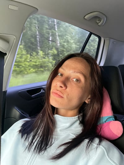 A female individual with medium-dark brown hair, brown eyes, and a tanned complexion with visible freckles is seated in the rear passenger seat of a vehicle, looking directly towards the camera. She has a gold-colored hoop piercing in her left nostril and a small gold stud piercing above her upper lip. Her lips are slightly pursed, and she is wearing a light blue, long-sleeved top with a zipper detail. A pink and light blue object, appearing to be a neck pillow or plush toy, is positioned behind her head against the black car seat. The vehicle's interior features a grey headliner with an "AIRBAG" label visible on the overhead panel to the left, a grey grab handle above the right rear window, and a black door panel with a silver handle on the left. Through the car windows, a blurred natural landscape of green trees and foliage is visible, indicating the vehicle is in motion. This scene is located in Pavlovka, Russia.