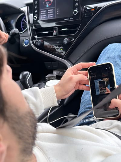 A person sitting in a car is holding a smartphone. The phone screen displays an image. The car's dashboard and steering wheel are visible. The location is not discernible.