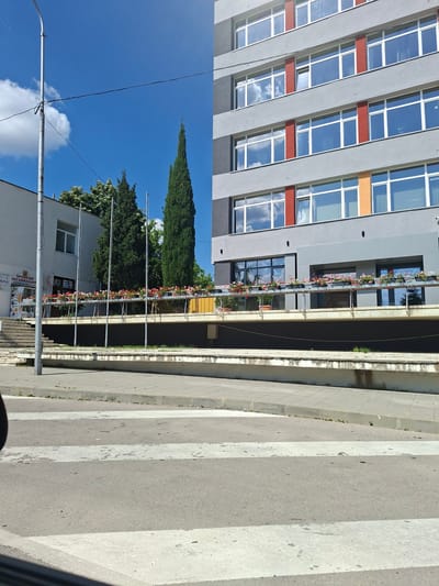 The image shows a street scene in Aksakovo, Bulgaria on a bright, sunny day. A modern, grey building with large windows and red and orange accents dominates the left side of the frame. The windows reflect a clear blue sky with white clouds. Flower boxes with red and pink flowers adorn balconies on the building.
To the right of the building, there is a pedestrian crossing with white stripes on a grey asphalt road. A sidewalk runs alongside the road, with a low concrete barrier separating it from a grassy area. In the background, a tall, conical evergreen tree stands prominently against the blue sky, along with other lush green trees. A white building with a window is visible behind the trees.
There are several metal poles on the right side of the image, one with a curved top, suggesting streetlights or traffic signals. Wires are strung between some of these poles.
Visible text on a sign on the white building reads "ПРЕВОДИ ЛЕГАЛИЗАЦИЯ". This translates to "Translations Legalization". Another sign on the same building is partially visible.
The overall impression is a clean and well-maintained urban environment, likely during daytime given the bright sunlight and clear sky. There are no people visible in the image.