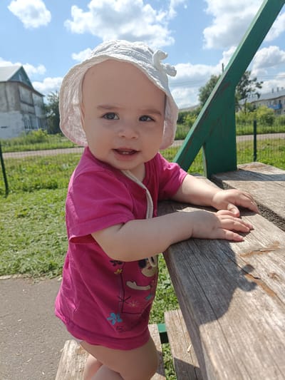 In a sunlit outdoor environment, a young child wearing a pink t-shirt and a white sun hat is positioned on a wooden structure. The child's face, with wide eyes and a slight smile, is visible. The child's hands are resting on the wooden surface. The background shows a residential building, a green fence, and green grass under a bright blue sky with scattered clouds. This scene is likely located in a residential area of Posad, Russia, possibly a park or a yard.