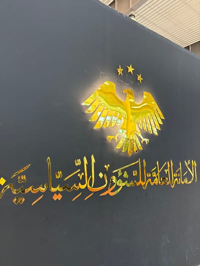 The image displays a dark, flat wall or panel with prominent golden, reflective signage, located in Al-Ghizlaniya, Syria. Centrally positioned is a stylized golden eagle emblem with outstretched wings, appearing to be backlit, creating a luminous halo effect. Above the eagle, three smaller golden stars are arranged in an arc. Below the eagle, golden Arabic calligraphy reads "الأمانة العامة للشؤون السياسية" (General Secretariat for Political Affairs). The reflective surfaces of the golden elements show distorted reflections of an unseen indoor environment, including what appears to be a light-colored, possibly ribbed, ceiling structure visible at the top edge of the frame. No individuals, actions, or direct interactions are depicted. The scene is static, focusing solely on the architectural signage.
