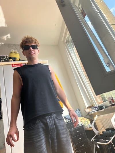 The image shows a young man with curly brown hair and sunglasses standing in a brightly lit room. He is wearing a black sleeveless shirt and dark wash jeans. To his left, on a shelf, is a stuffed Minion toy and a red "Supreme" sticker. Sunlight streams in from a large window on the right side of the frame, illuminating the scene. The room appears to be a residential interior. The city of Hamm, Germany, is not depicted or referenced in this image.