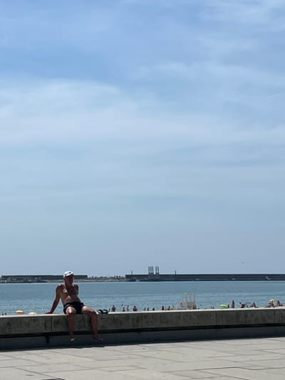 The image depicts a man sitting on a low concrete wall overlooking the ocean. He is wearing black shorts and a white cap, with his torso exposed. His legs are dangling over the edge of the wall. In the background, the ocean stretches to the horizon, with a long, dark structure, possibly a breakwater, running across the water. Several other people are visible on the beach behind the wall, some lying down, others standing or walking. A lifeguard station is also present on the beach. The sky is clear and blue, with some faint clouds. The scene appears to be set in a public beach area, likely during the daytime. The composition and presence of the breakwater suggest a coastal urban environment. Given the provided location of Matosinhos, Portugal, this scene could be representative of a typical summer day at one of its beaches.