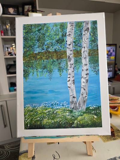 The image displays an acrylic painting on canvas, leaning against a wooden easel. The painting depicts a natural scene with a focus on birch trees. Two prominent, thick birch tree trunks with their characteristic white and black markings run diagonally across the canvas from the bottom left towards the top right. To the left of the birch trees, a dense area of green foliage, suggesting more trees or dense undergrowth, is painted with varying shades of green and hints of brown and yellow. To the right of the birch trees, a vibrant field of green grass is depicted, dotted with small white flowers. The background is a clear, bright blue sky. The style of painting is impressionistic, with visible brushstrokes adding texture to the foliage and grass.
The painting is set up on an easel, suggesting it might be in a home or studio environment, possibly in Claremorris, Ireland, as per the location context. The lighting suggests daytime, with the bright blue sky indicating fair weather. No specific activity or event is depicted, as the focus is on the landscape. There are no people visible in the painting or immediately around it. No text is visible on the painting itself. The overall impression is a serene and verdant natural landscape.