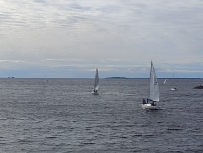 The image captures a cloudy day at sea, with several sailboats visible. In the foreground, a white sailboat with sails up is sailing from right to left. It has three people on board, two of whom are standing. The number "FIN 9069" is visible on its sail. Further in the distance, another white sailboat is also sailing. To the far right of the image, a rocky outcrop is visible with a few people standing on it. The water is choppy and dark grey. The horizon is lined with a faint landmass under a sky filled with layered, grey clouds. The scene is likely the Bay of Bothnia, near Oulu, Finland, possibly at Nallikari beach, given the presence of sailboats and the general coastal landscape.