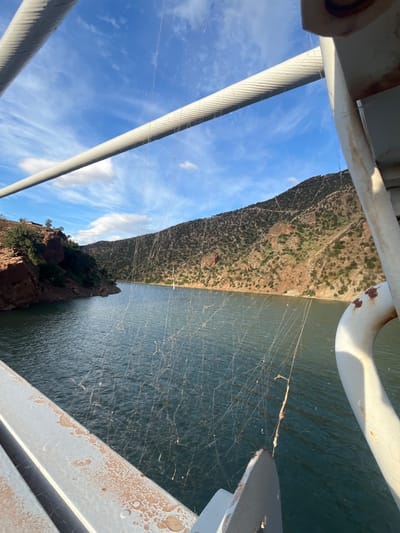 The image shows a view from a bridge spanning a body of water adjacent to a mountainous landscape. The bridge structure is visible in the foreground, with large cables and white-painted railings. The water appears dark and possibly still, with numerous cobwebs visible. The landscape on the other side of the water consists of a steep, rugged hill covered in sparse vegetation. The sky above is blue with scattered clouds. There is no evidence to suggest this scene is in the city of Ait Mazigh, Morocco.