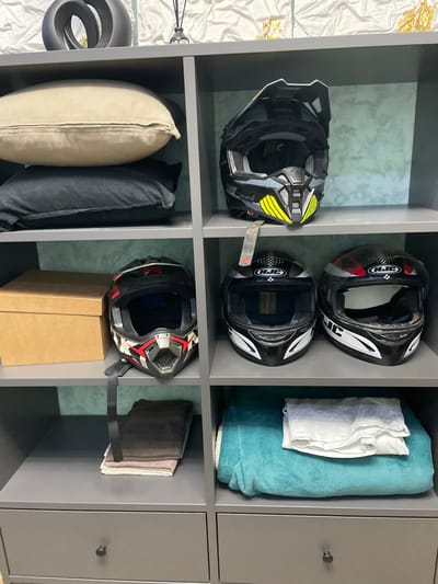 The image depicts a gray shelving unit or closet containing various items. On the top left shelf, there is a black motocross helmet with yellow accents. Next to it, on the top middle shelf, there are two black motorcycle helmets with white and red details. The top right shelf contains a folded light blue towel and a white folded shirt or cloth. The bottom left shelf has a beige pillow and a black pillow. Below the middle shelf, there is a black motorcycle helmet with red and white accents sitting atop a brown cardboard box. On the bottom right shelf, there is a folded brown towel. The shelves are part of a modular storage system with a door on the rightmost section. There are no people visible in the image. The location, Лесной Пейзаж Кп (шарапово), Russia, is not visually represented in the image.