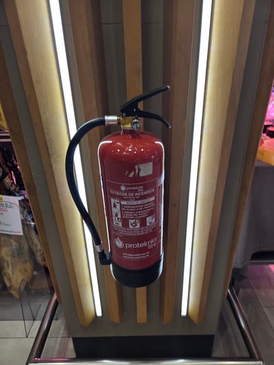 A red fire extinguisher is mounted on a wooden structure with vertical panels and integrated lighting. The fire extinguisher has a black hose and a silver nozzle. It is a Proteknia brand, model "EXTINTOR DE INCENDIOS". The label indicates it is for ABC powder, 6kg, and suitable for use on electrical fires up to 50kV at a minimum distance of 1 meter. The fire extinguisher is positioned in a commercial setting, with a display of cured meats visible in the background to the left. To the right, there is a glimpse of a light-colored fabric and some black cylindrical objects. The overall environment suggests a public space like a market or a store. The provided crops offer closer views of the fire extinguisher's label and the surrounding wooden structure.