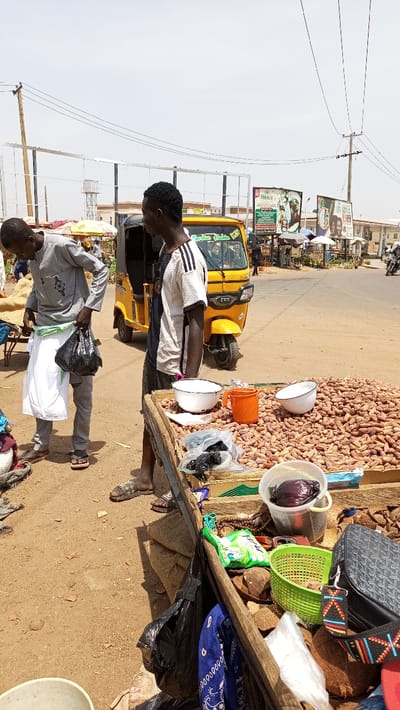 The image depicts an outdoor market scene in Barnawa, Nigeria, under bright daylight. In the immediate foreground, a makeshift wooden stall is laden with various goods. A large quantity of light brown, elongated produce, consistent with dried dates or groundnuts, occupies the main surface. Alongside these, white enamel bowls, an orange plastic cup, several clear plastic bags containing dark items, a green plastic basket, and a clear bucket holding a dark red cabbage are arranged. Other items on the stall include coconut halves, a patterned shoulder bag, a blue bandana, brown beads, and a small packaged item.

Two adult males are prominent in the foreground. On the left, a man wearing a light grey tunic with black embroidery, grey trousers, and brown sandals is partially bent over. He holds a black plastic bag in his right hand and appears to be adjusting a white cloth around his waist with his left hand. To his right, another man stands upright, wearing a white and black striped short-sleeved t-shirt, dark trousers, and dark patterned slide sandals. He faces left, away from the viewer.

Behind the individuals and the stall, a paved road is visible. A yellow TVS-branded auto-rickshaw (Keke Napep) with "Garden Hukba KFC" text is positioned on the road, facing left. The surrounding environment includes dry, light brown earth transitioning to the asphalt road. In the midground and background, utility poles with overhead wires are present. Multiple billboards are visible, including one advertising "Pukka's" and another with "Garden Hukba KFC" branding. Various buildings, distant market stalls with umbrellas, and a few individuals, including a person on a motorcycle, are discernible further down the road. The sky is clear and light-colored, indicating a sunny condition.