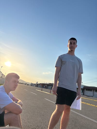 A photograph shows a man standing on an asphalt surface in the city of Dimitrovgrad Municipality, Serbia. The man is wearing a light-gray t-shirt and black shorts, and he is holding sheets of paper in his hands. The asphalt appears to be a road or parking area, marked with white lines. The background includes buildings, utility poles, and a clear sky with the sun low on the horizon, suggesting either dawn or dusk. A second person is seen in the foreground, seated with their legs crossed, wearing a white shirt.