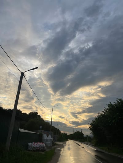 The image depicts a wet, empty road in Солнечная Поляна, Russia, under a dramatic, cloudy sky with shafts of sunlight breaking through. The road surface is reflective, indicating recent rain. On the left side of the road, a concrete utility pole with a street lamp and electrical wires stands prominently. Adjacent to the pole is a green corrugated metal fence. Piles of white and red-bagged materials, possibly construction supplies, are stacked on pallets near the fence. Further down the road on the left, there's a bus stop sign. On the right side of the road, dense green trees line the edge. In the distance, more trees and what appear to be utility poles are visible. The overall scene suggests a rural or semi-rural setting at either dawn or dusk. There are no people or animals visible in the image.