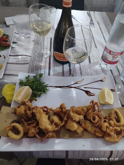 This media file depicts a close-up, overhead shot of a table setting at a restaurant. The main subject is a plate of golden-brown, crispy fried calamari served on a brown paper liner atop a white rectangular plate. The calamari appears to be a mix of rings and tentacles. Accompanying the calamari are two lemon wedges, one on the plate and one partially visible to the left. A small pile of fresh green arugula is also present on the plate.
In the background, two wine glasses filled with a pale yellow liquid, presumably white wine, are visible. Between the glasses, a dark green bottle with a distinctive orange and white label is placed. The label appears to have the word "CHARME" prominently displayed. To the right of the glasses, a clear plastic bottle of water with a red and white label is also present. The table is covered with a light-colored wooden surface with a grain pattern. The setting suggests a meal is being enjoyed, likely during the evening as indicated by the time stamp. The overall mood appears relaxed and convivial.
Visible text includes "CHARME" on the wine bottle and "26/06/2026 21:37" as a timestamp in the lower right corner. The location context is Scordia, Italy.