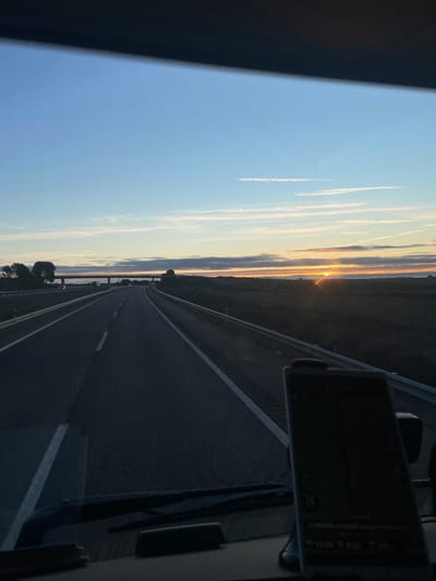 A highway stretches into the distance, illuminated by a setting sun. The road is bordered by fields and trees. Jet contrails mark the blue and orange sky. A navigation device is mounted on a dashboard to the right of the view, showing map details and the time. The photo is taken from the interior of a vehicle looking forward. The image is rotated.