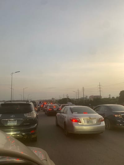 The image depicts a congested highway with multiple lanes of stationary vehicles during what appears to be rush hour. The perspective is from a vehicle looking forward, capturing a long line of cars, SUVs, and trucks. Overhead streetlights are visible along the side of the road, and in the distance, power lines and buildings can be seen. The sky is overcast, with the sun setting or rising, casting a warm, diffused light. The scene is characterized by the stillness of traffic, with brake lights illuminating the road ahead. There are no visible people outside of the vehicles. The location is identified as Kuchingoro, Nigeria.