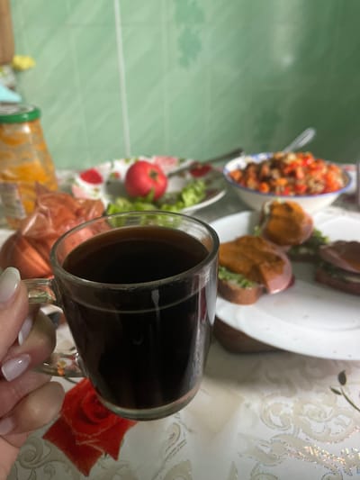 A close-up shot captures a clear glass mug filled with dark coffee held by a person's hand with light-colored nails. In the background, a table is laden with food items. To the left, a glass jar with an orange substance and a transparent plastic bag containing a few items are visible. Further back, a plate with a red tomato and green lettuce is present, along with a bowl of salad. To the right, a white plate holds several open-faced sandwiches topped with what appears to be meat and sauce, with one sandwich in focus. The setting suggests a meal in a kitchen or dining area, likely within a home in Красное, Russia. The overall atmosphere is casual and domestic.