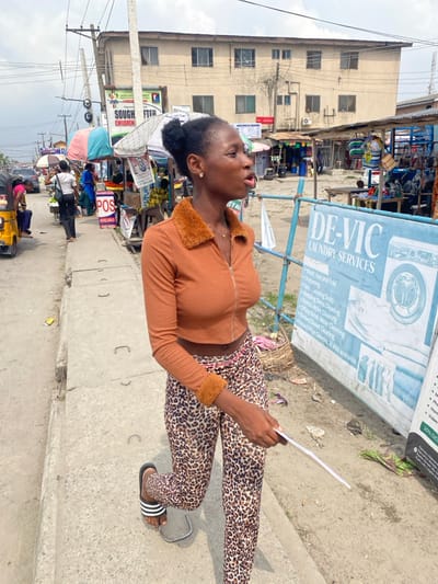 The image depicts a daytime street scene in Langbasa, Nigeria, characterized by a bustling urban market environment. In the foreground, a young Black female, with dark skin and her black hair styled in a bun, walks from right to left on a concrete sidewalk. She is wearing an orange ribbed crop top with a brown faux-fur collar and cuffs, leopard-print leggings, and black Adidas slides. A navel piercing is visible. She holds a white paper or leaflet in her right hand, her mouth slightly open, suggesting she is speaking or exhaling. Her gaze is directed towards the left.

To her right, a large blue and white sign for "DE-VIC LAUNDRY SERVICES" is prominently displayed, listing services such as "Wash, Iron and Fold," "Steaming," "Dry Cleaning," and "Wedding Dress Cleaning." Beyond the sign, a dirt path separates the sidewalk from several makeshift market stalls constructed with metal frames and various tarpaulins, displaying goods. A multi-story, light-colored building with multiple windows stands in the background.

To the left of the main subject, the concrete sidewalk continues alongside a busy asphalt road with vehicles, including a yellow auto-rickshaw (keke Napep), and multiple pedestrians. Numerous market stalls with colorful umbrellas and signs, some partially obscured, such as "SOUGHT AFTER" and "POS," line the street. Overhead utility poles and a dense network of power lines are visible against an overcast sky. People are seen walking, standing, and engaging in market activities throughout the midground.