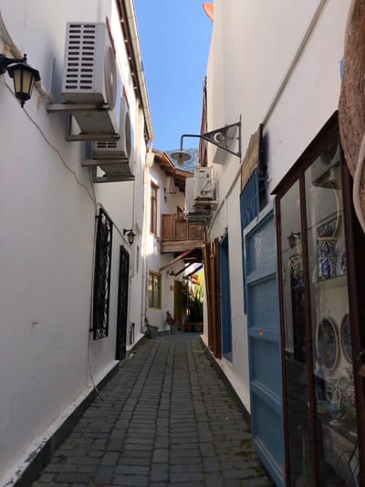 The image depicts a narrow cobblestone alleyway in Kaş, Turkey. The alley is flanked by white-washed buildings with traditional architecture. On the left side of the alley, an outdoor lamp and two air conditioning units are mounted on the wall. Further down the alley on the left, there are two windows with ornate black wrought iron grills. On the right side of the alley, there is a shop with a glass display case filled with decorative pottery and plates. A blue wooden door is also visible on the right. The alley is empty of people. The sky is clear and blue. The overall impression is one of a quaint and charming Mediterranean street.