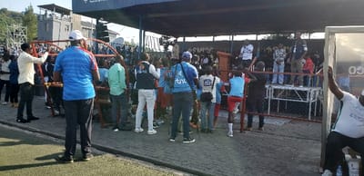 A crowd of people gathers near a podium under a covered area. Some people are standing behind a red metal fence, observing the event. Several individuals are wearing vests with logos, possibly indicating they are part of event staff or media. A few are standing on a metal platform for a better view. Some are dressed in casual clothes. The overall scene suggests a public event, possibly a speech or presentation.
