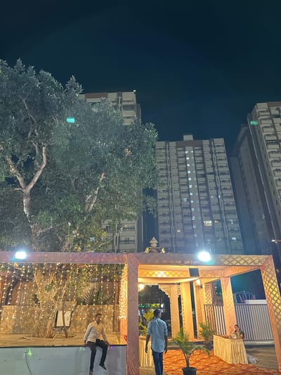 This nighttime shot, taken in Nashik, India, features a decorated outdoor event space. An elaborately decorated awning, lit with strings of lights, dominates the right side of the frame. A man in a blue shirt stands partially under the awning, while another man in a striped shirt walks near a wall behind the awning. Behind the event space, a large tree fills the center of the image. In the background, tall apartment buildings rise into the night sky.
