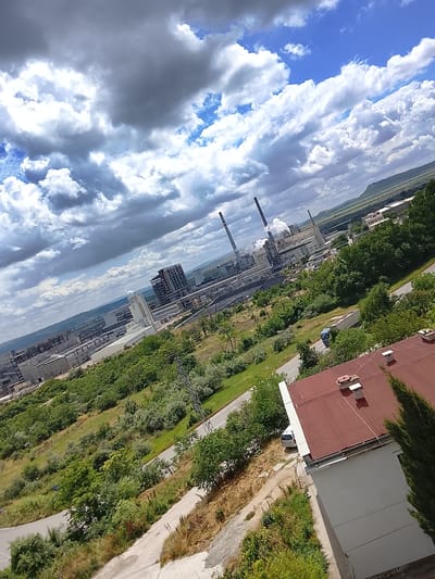 The image depicts an industrial area in Devnya, Bulgaria, on a cloudy day. In the foreground, a portion of a building with a reddish-brown roof and a white wall is visible. Adjacent to this building, a tall, dark green coniferous tree stands on the right.
The middle ground of the image shows a winding road with a light-colored car parked on the side. Surrounding the road are green trees and shrubs, with a power line tower visible in the distance.
The background features a large industrial complex with multiple buildings and smokestacks emitting plumes of smoke. The complex appears to be a thermal power plant or similar facility. Beyond the industrial zone, rolling hills covered in vegetation are visible under a dramatic sky filled with heavy, dark clouds interspersed with patches of blue. The overall scene suggests an active industrial operation within a naturally scenic landscape.