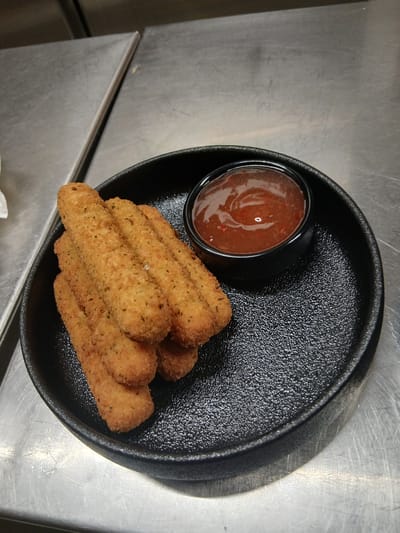 The image depicts a meal consisting of fried breaded sticks and a dipping sauce, presented on a black serving dish. This scene is situated on a metallic countertop, typical of a commercial kitchen environment in Sotira, Cyprus.

Specifically, approximately six to seven golden-brown, cylindrical, breaded food items, consistent with mozzarella sticks, are stacked centrally on a round, black, textured plate. The breading on these items features visible green seasoning flecks. To the right of the food items, a small, black, cylindrical ramekin contains a reddish-orange, viscous dipping sauce, which appears to be sweet chili sauce due to visible red chili flakes and its glossy surface reflecting ambient light. The black plate has a distinct pebbled surface texture and a raised rim.

The entire serving arrangement rests on a brushed or scratched stainless steel countertop. A vertical seam or division in the metallic surface is visible in the upper left background. No individuals or further human actions are depicted.