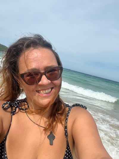 A woman is taking a selfie on a beach in Cam Lâm, Vietnam. She is wearing a black and white polka dot bikini top with ties on the shoulders and a dark pendant necklace. She has sunglasses on and is smiling. The ocean is visible behind her with waves breaking on the shore. The sky is clear and blue.