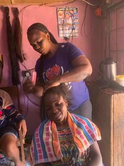The image captures an indoor scene in Garaku, Nigeria, depicting a hair styling activity within what appears to be a small salon or a home-based styling area. A dark-skinned adult female, the stylist, stands wearing a purple short-sleeved t-shirt and grey bottoms, her hair in braids. She is actively working on the hair of a seated dark-skinned adult female client. The stylist holds a pink comb in her left hand, with both hands manipulating the client's natural, unbraided hair. The client wears a multi-colored striped towel draped over her shoulders and a patterned top, looking downwards with a slight smile. A third individual is partially visible on the far left, seated on a wooden chair, wearing a patterned top and shorts.
The room features light pink walls. To the left of the stylist, two bundles of dark hair extensions hang. A black oscillating fan is positioned behind the stylist. On a wooden surface to the right, a silver electric kettle, a yellow lidded jar, and black hair styling tools including a flat iron and combs are arranged. A calendar-style poster titled "HAPPY MARRIED LIFE." with multiple images is affixed to the wall, above which exposed electrical wiring is visible. Natural light enters the space, casting highlights and shadows.